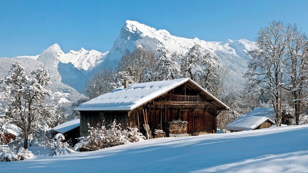 Eine Holzhütte mit schneebedecktem Dach, umgeben von schneebedeckten Bäumen und Bergen im Hintergrund.