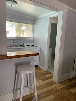 Une cuisine avec un plan de travail en bois, un évier blanc et une cuisinière. On y trouve un tabouret blanc et une fenêtre avec des stores.