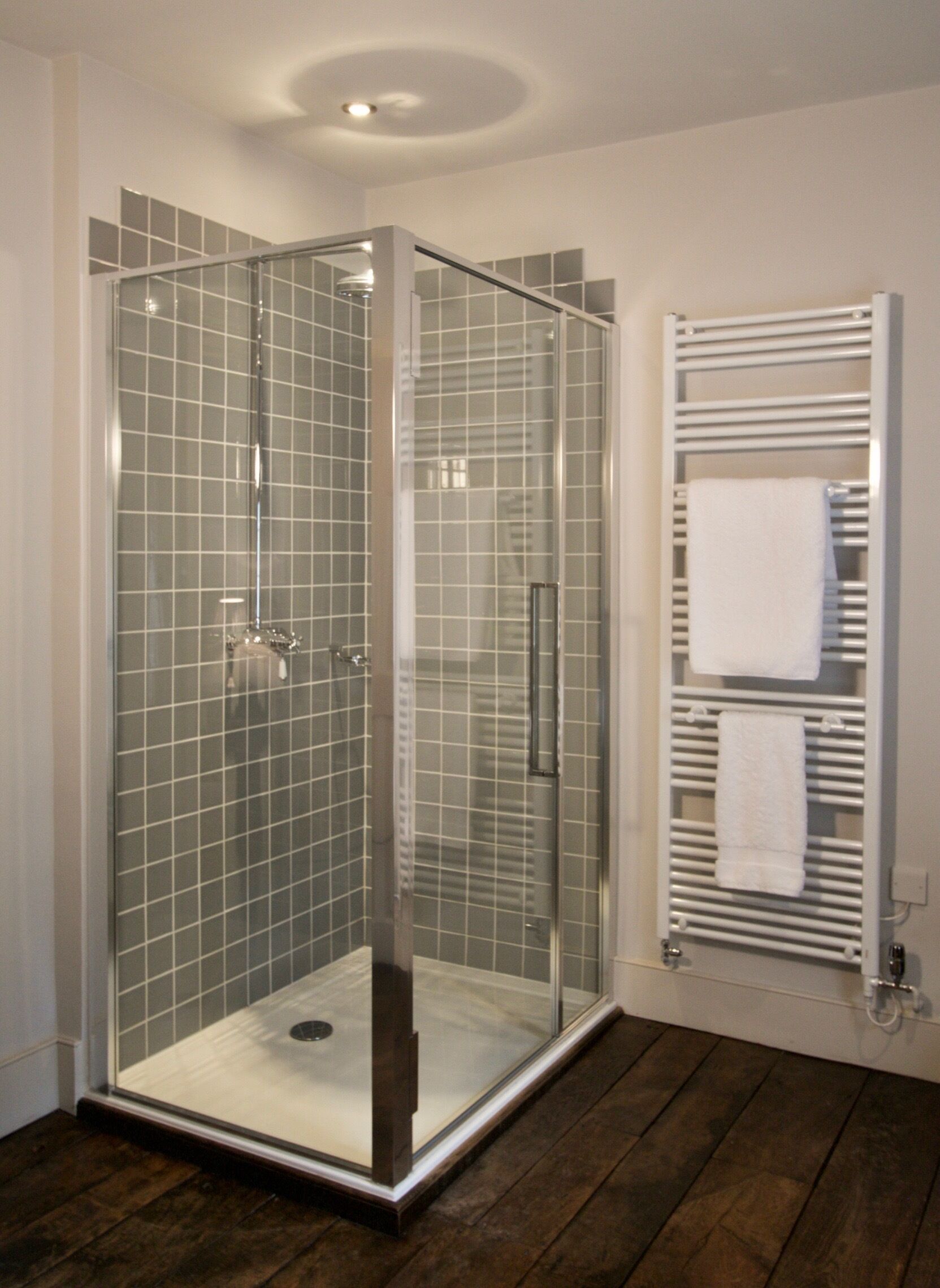 A glass-enclosed shower with a tiled floor, a white towel rack, and a wooden floor.