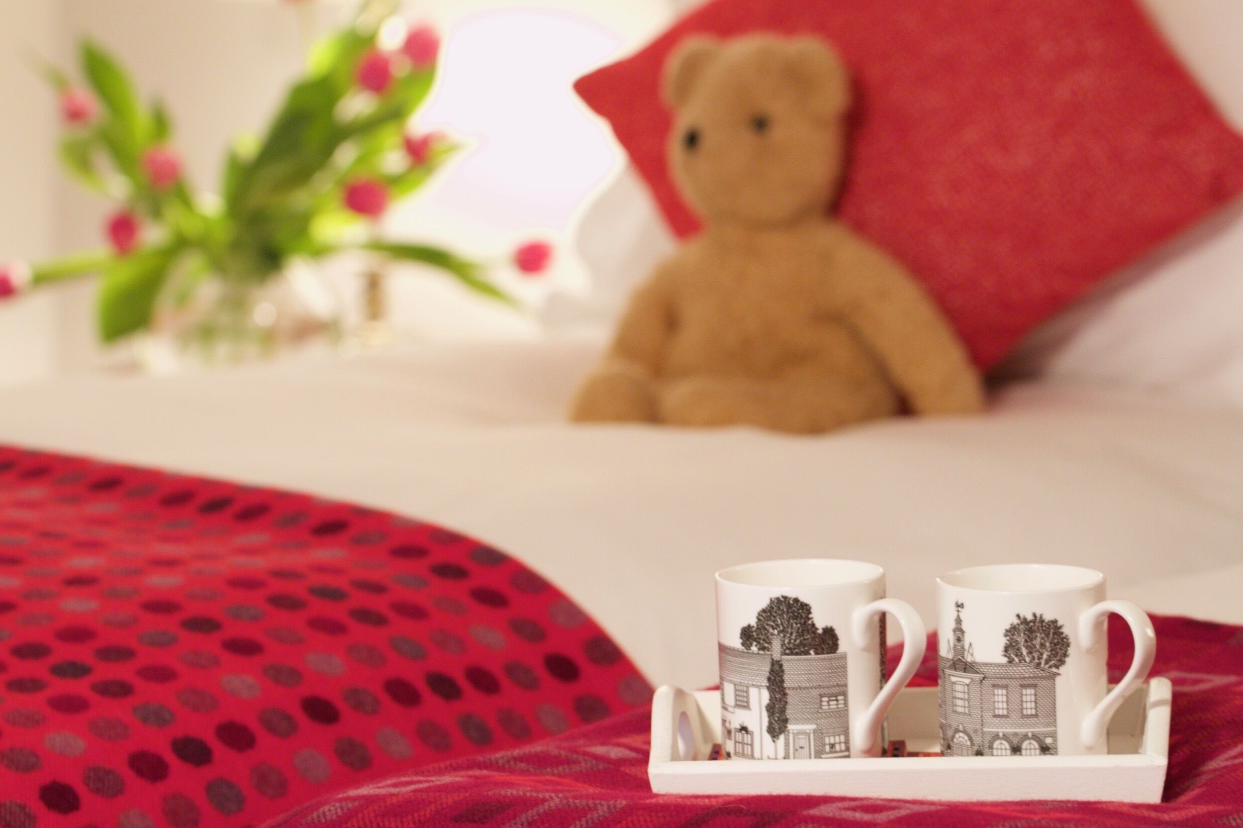 A bed with a red polka dot blanket, a teddy bear, and a tray with two mugs featuring a building design.