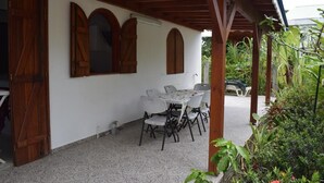 Um pátio externo com mesa e cadeiras, rodeado por vegetação e uma área coberta com vigas de madeira.