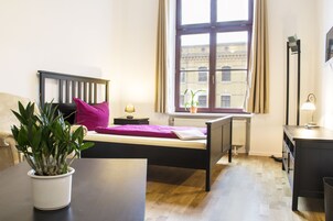 A bedroom with a bed, bedside table, lamp, potted plant, and a window with a view of buildings.