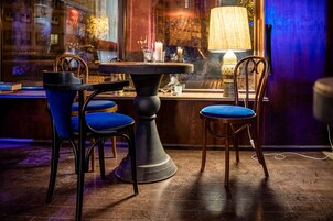 A bar area with a round table, two blue cushioned chairs, a lamp, and a window with reflections of the city.