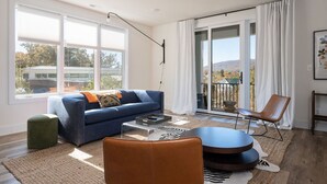 A modern living room with a blue sofa, a leather armchair, and a coffee table. There is a large window with a view of trees and a building outside.