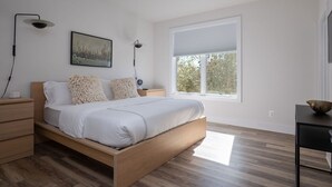 A modern bedroom with a wooden bed, white bedding, a large window with blinds, and a nightstand with a lamp.