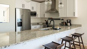 A modern kitchen with white cabinets, a stainless steel refrigerator, a built-in oven, a microwave, a gas stove, and a coffee maker. There are two bar stools and a hanging light fixture.