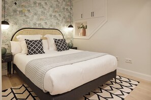 A neatly made bed with a patterned throw, a bedside table with a lamp, and a wall with a floral wallpaper and a shelf with a plant. - Hygge by FeelFree Rentals (Donostia)