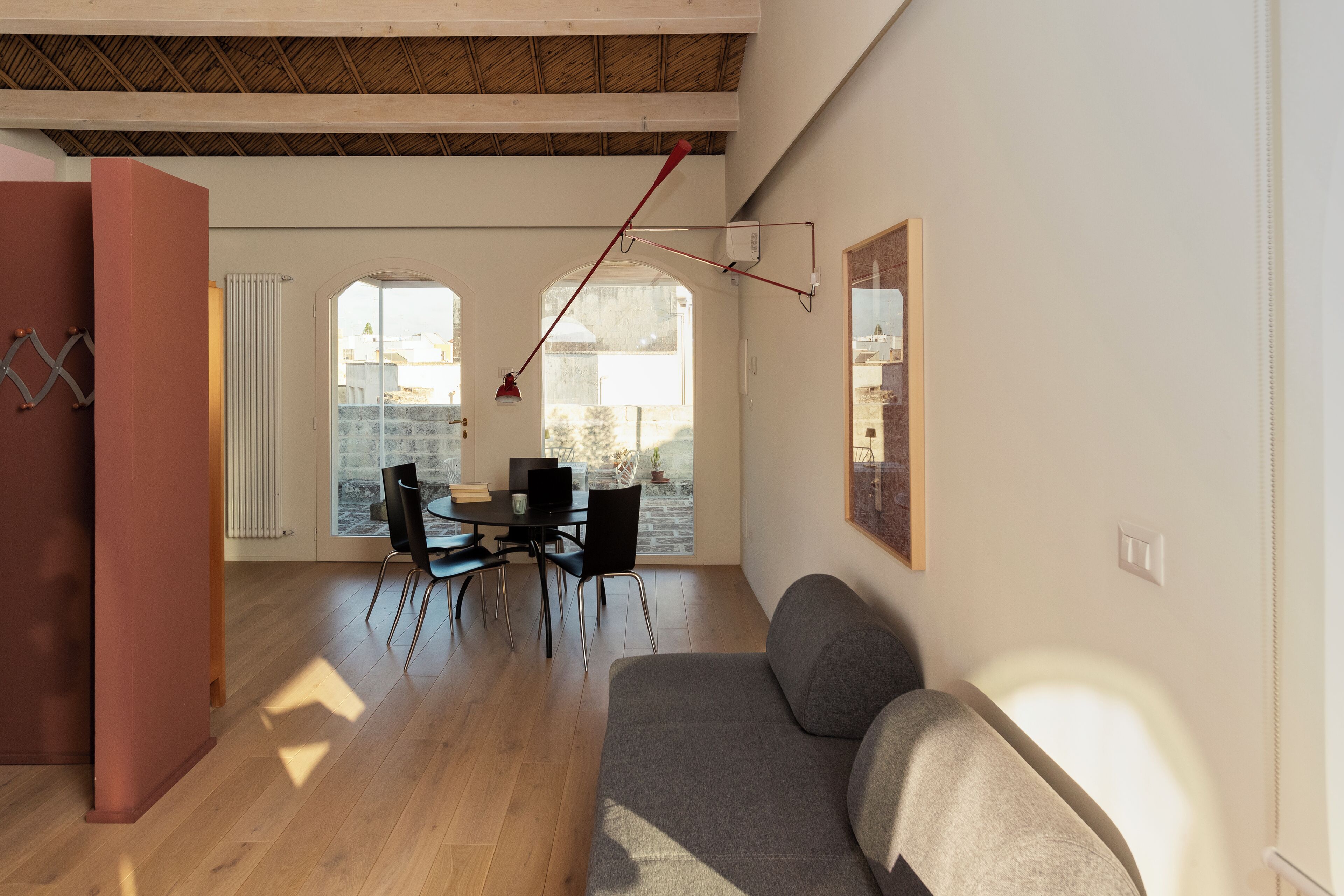 A modern living room with a sofa, dining table, and chairs. There is a large window providing natural light and a view of the outside.