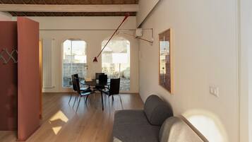 A modern living room with a sofa, dining table, and chairs. There is a large window providing natural light and a view of the outside.