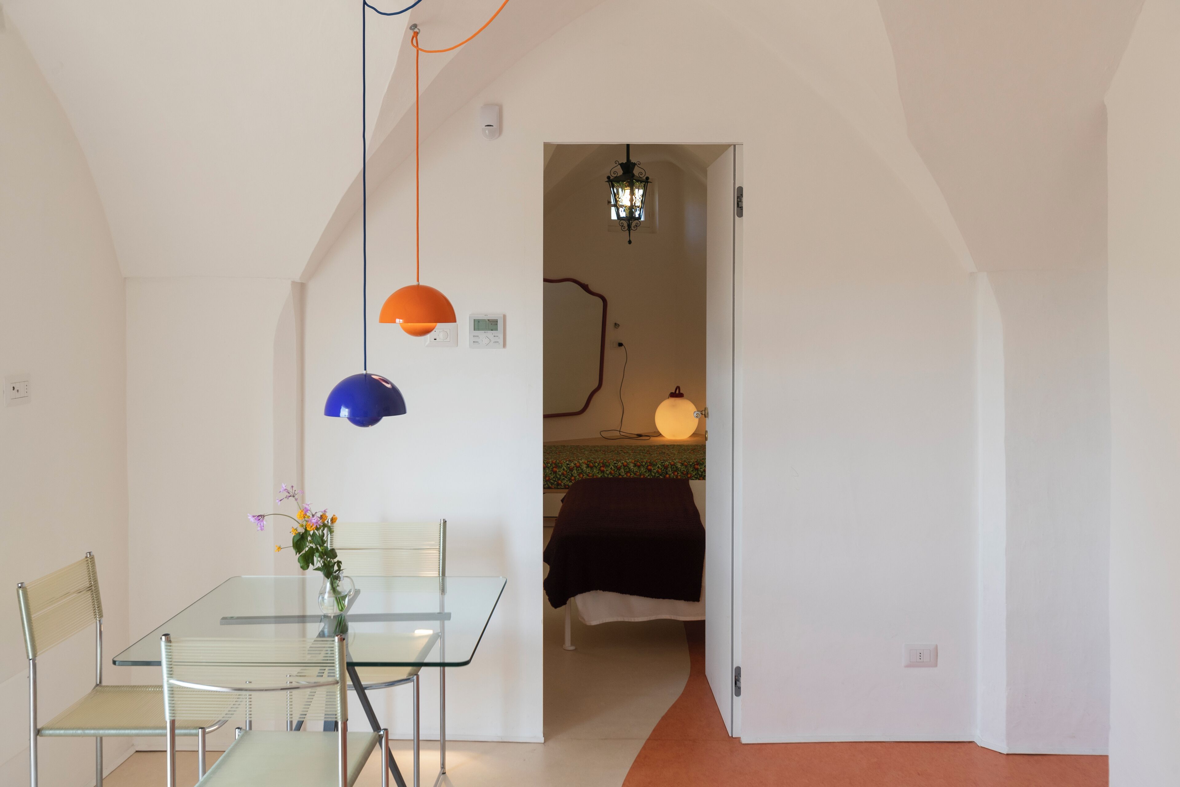 A modern room with a glass table, metal chairs, a bed with a black cover, and a hallway leading to another room.