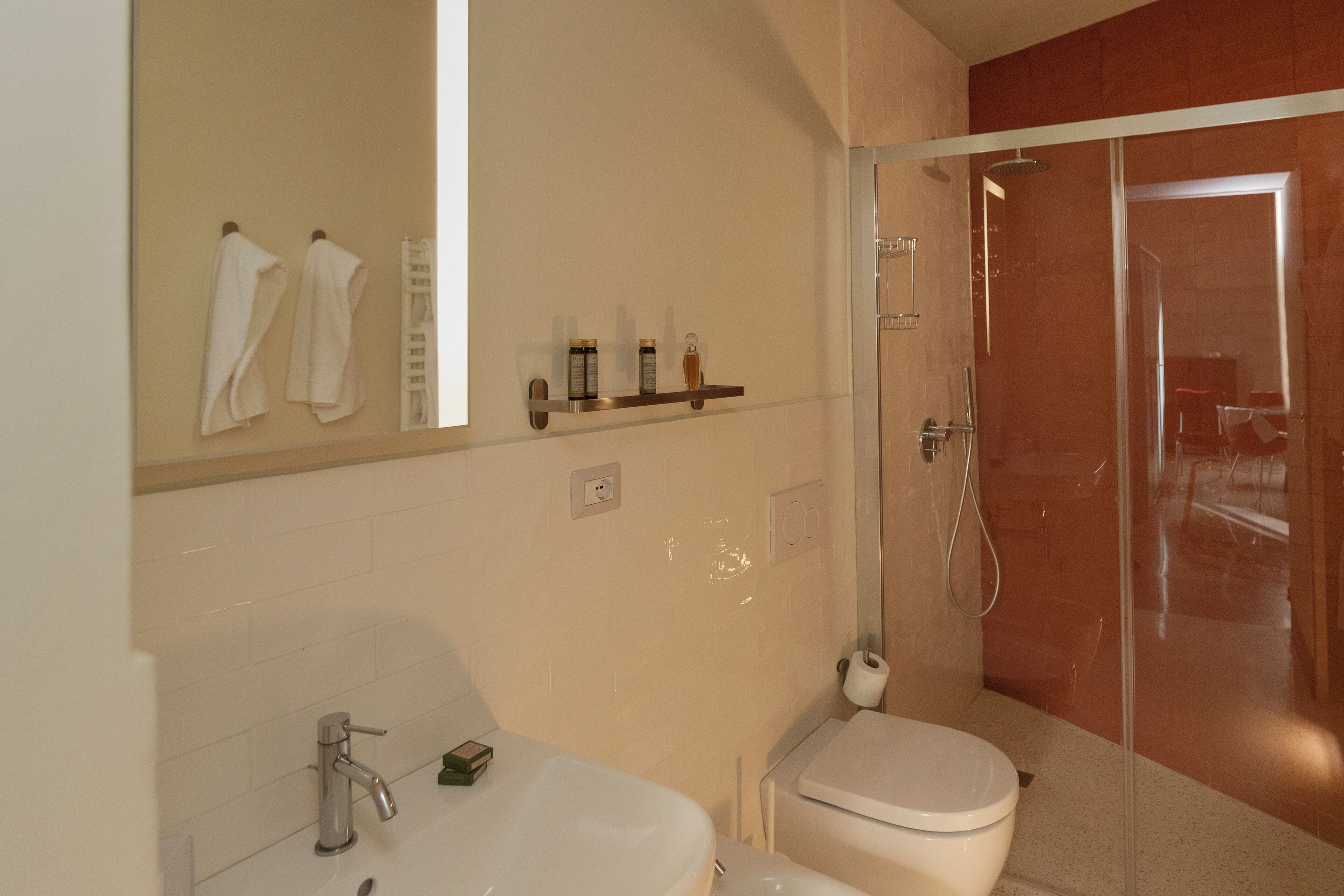 A bathroom with a sink, toilet, and a glass-enclosed shower. There are towels hanging on the wall and a shelf with toiletries.