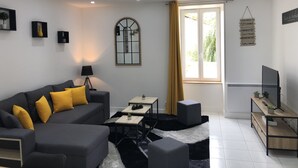 Una sala de estar moderna con un sofá gris, cojines amarillos, una mesita con plantas, un mueble para la televisión y una ventana con cortinas.