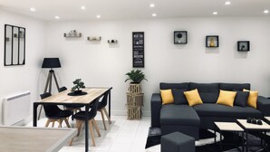 Una sala de estar moderna con un sofá negro, una mesa de comedor de madera y sillas negras. Hay una lámpara de pie negra, una planta en maceta y un cuadro en la pared.