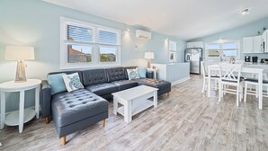 A modern living room with a sectional sofa, a dining table with chairs, and a kitchen area. - The Surf  - Surf's Up!  Oceanfront  Getaway!!   (North Topsail)