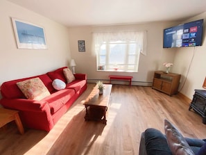Un salón con un sofá rojo, una mesa de centro de madera, un banco, un televisor montado en la pared y una ventana con persianas.