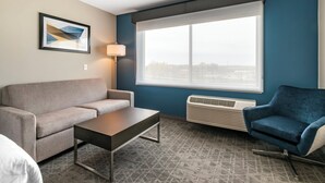 Een hotelkamer met een grijze bank, een houten bijzettafeltje, een blauwe fauteuil, een groot raam met uitzicht en een wandairconditioner.