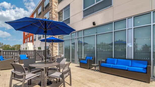 Een terras buiten met een tafel, stoelen, blauwe parasols en een bank met blauwe kussens.