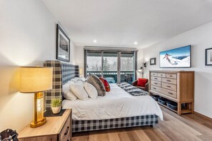 A modern bedroom with a bed, bedside tables, a dresser, and a large window with a view of trees and mountains. - Snowloft Ski In & Ski out (Beaver Creek)