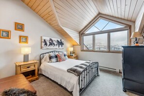 A bedroom with a sloped ceiling, a large window, a bed, bedside tables, and a dresser. - Snowloft Ski In & Ski out (Beaver Creek)