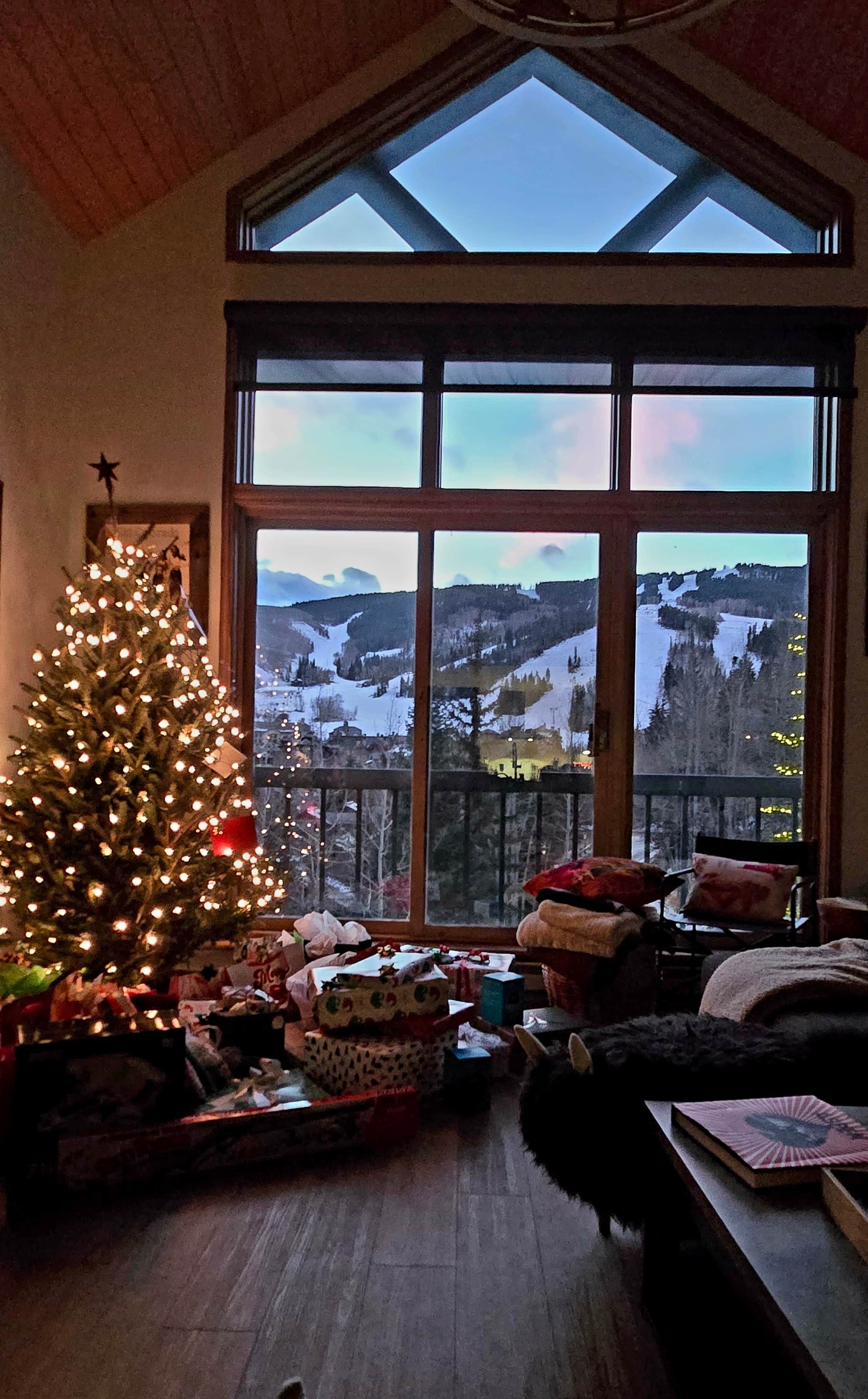 Une pièce dotée d’une grande fenêtre donnant sur un paysage enneigé, d’un sapin de Noël décoré et d’un coin salon confortable.