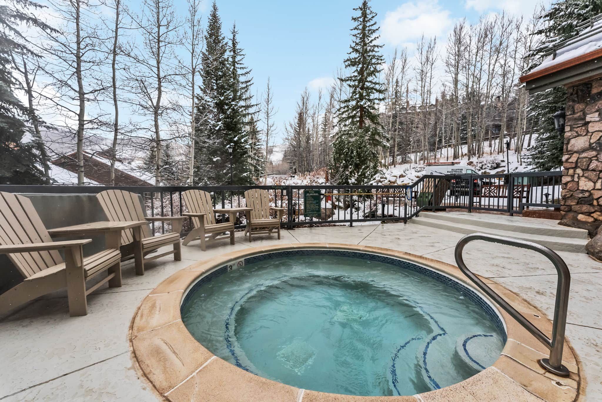 A hot tub surrounded by Adirondack chairs, a stone building, and a snowy forest backdrop.