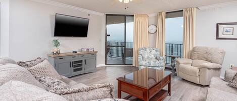 A spacious living room with a large sofa, a coffee table, a TV mounted on the wall, a ceiling fan, and a view of the ocean through the sliding glass doors.