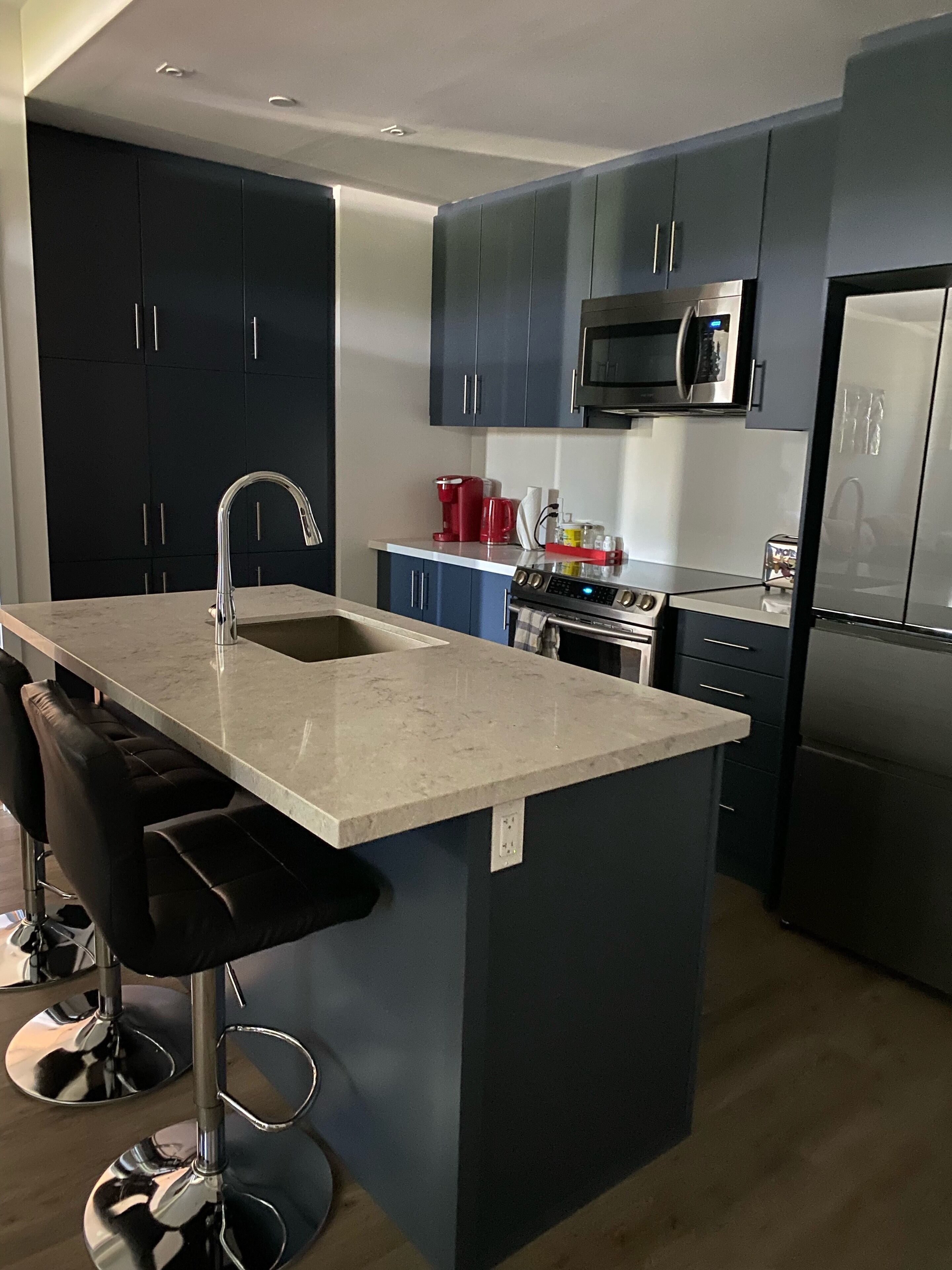 A modern kitchen with dark cabinets, stainless steel appliances, a marble countertop, and bar stools.