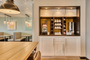 A restaurant with a wooden table, cushioned seating, pendant lighting, and a coffee station with various cups and a coffee machine. - Premier Inn Manchester Cc Princess Street (Manchester)