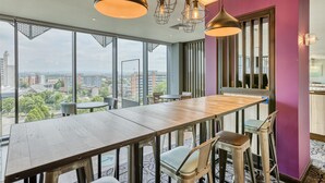 A modern dining area with a long wooden table, bar stools, and pendant lighting. - Premier Inn Manchester Cc Princess Street (Manchester)