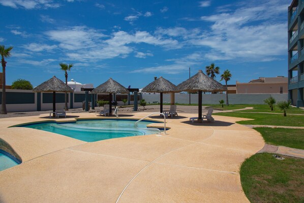 A pool area with lounge chairs and thatched umbrellas, surrounded by palm trees and a clear sky. - Linda Vista 105 (2Bedroom-2Bathroom) By FMI Rentals (Puerto Peñasco)