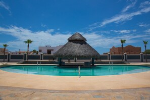 A swimming pool with a thatched-roof structure in the center, surrounded by palm trees and buildings. - Linda Vista 105 (2Bedroom-2Bathroom) By FMI Rentals (Puerto Peñasco)