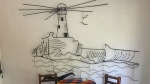 Een eetgedeelte met een tafel bedekt met een kleurrijke tafelkleed, twee stoelen en een muurschildering van een vuurtoren en een schip.