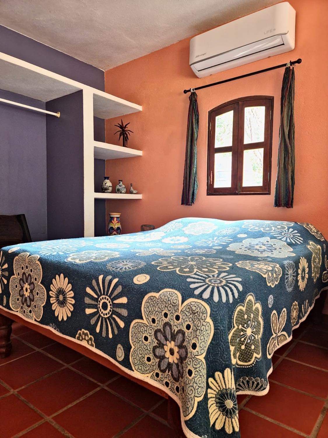 Une chambre à coucher avec un lit, une fenêtre avec des rideaux, une étagère avec des objets décoratifs et un appareil de climatisation.