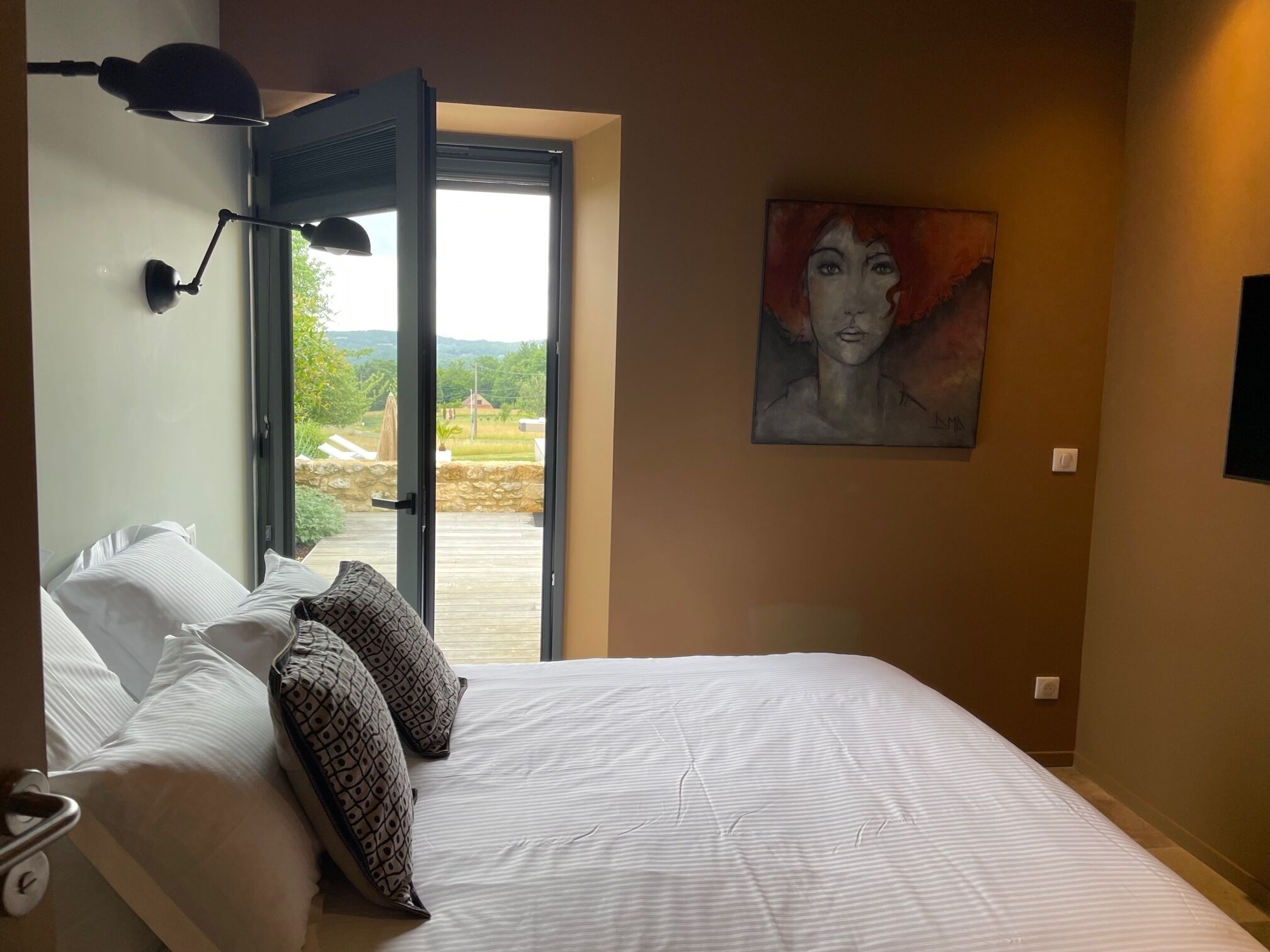 Une chambre à coucher avec un lit, des oreillers, une lampe de bureau, un tableau et une porte ouverte donnant sur un balcon avec vue sur la verdure.