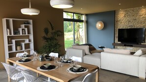 A modern living room with a dining area, a TV mounted on a textured wall, and a large window with a view of greenery.