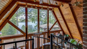 Eine Hütte mit einem großen Erkerfenster, das Blick auf einen See und einen Wald bietet. Eine Treppe führt zu einer oberen Terrasse mit Sitzgelegenheiten im Freien.