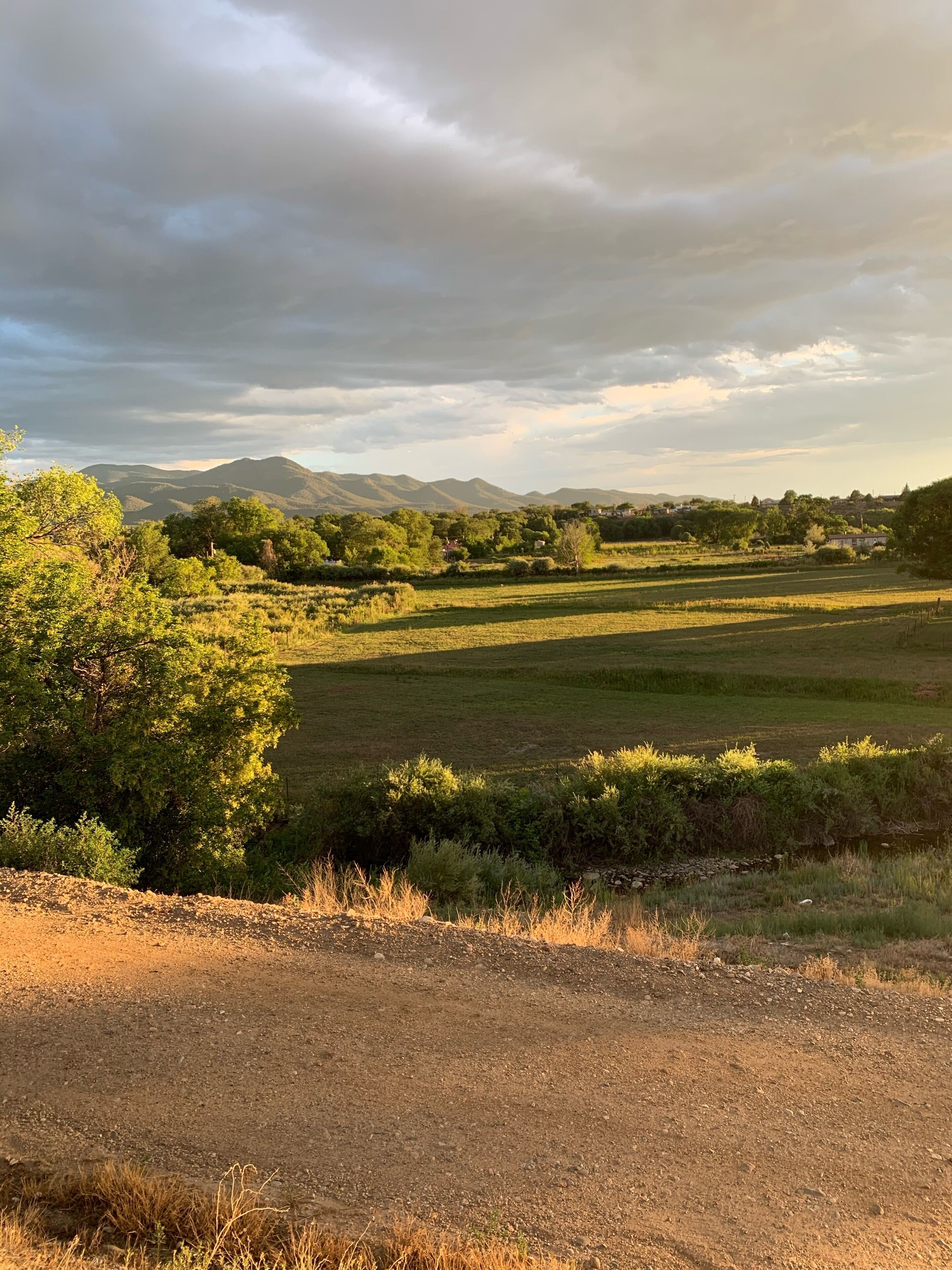 A rural landscape with fields, trees, and distant mountains under a cloudy sky.