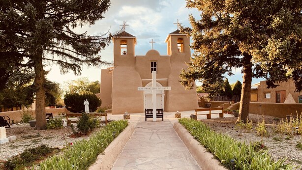 A church with a central entrance, flanked by two towers, surrounded by a garden with a pathway leading to the entrance.