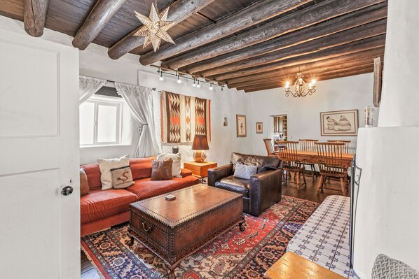 A cozy living room with a red sofa, a leather armchair, a wooden coffee table, and a patterned rug. There is a dining area with wooden chairs and a table, and a chandelier hanging from the ceiling. - Taos Hacienda Del Sol (Ranchos de Taos)