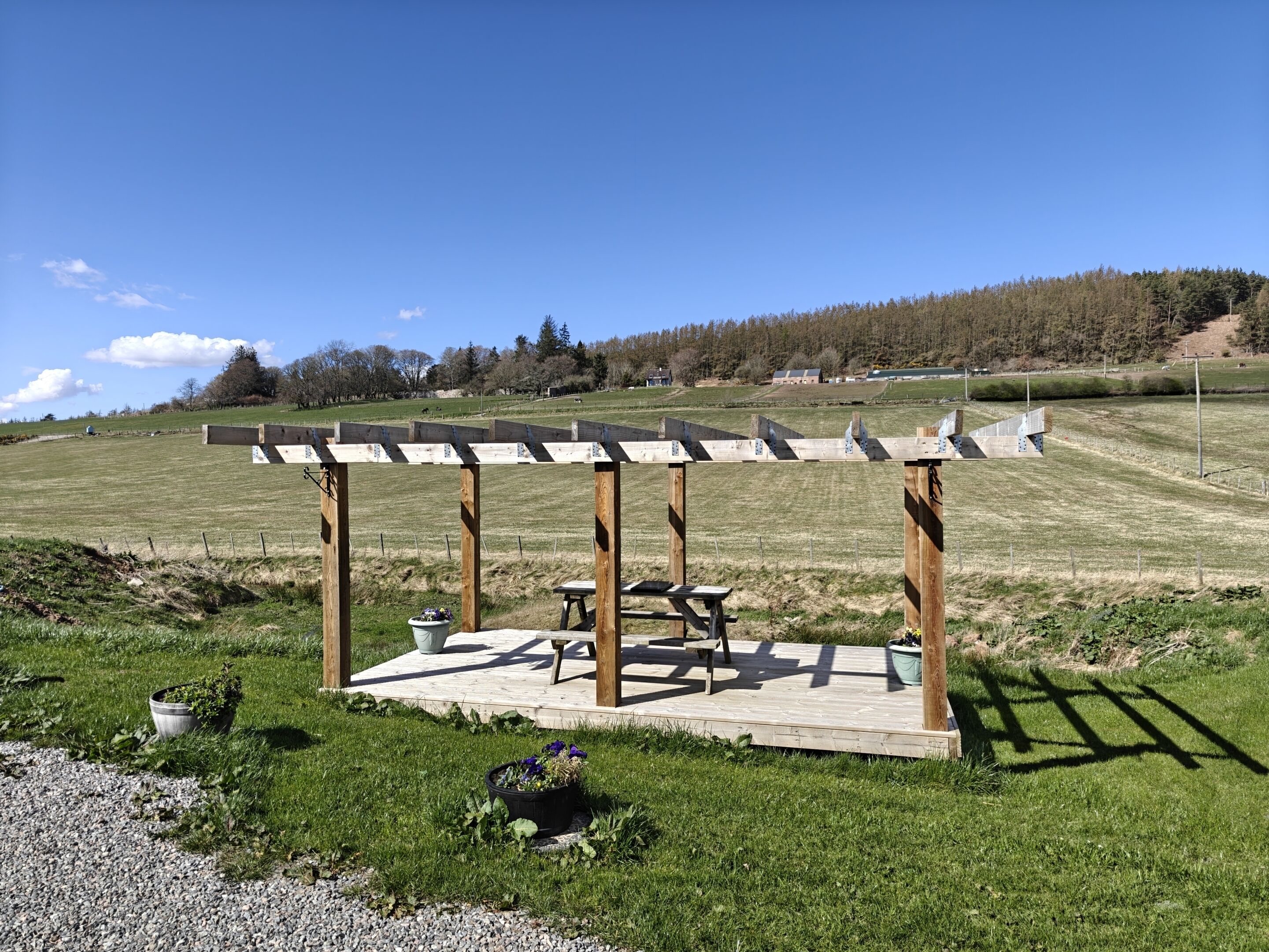Une pergola en bois, avec une table et des chaises, entourée d’un champ herbeux et d’arbres.