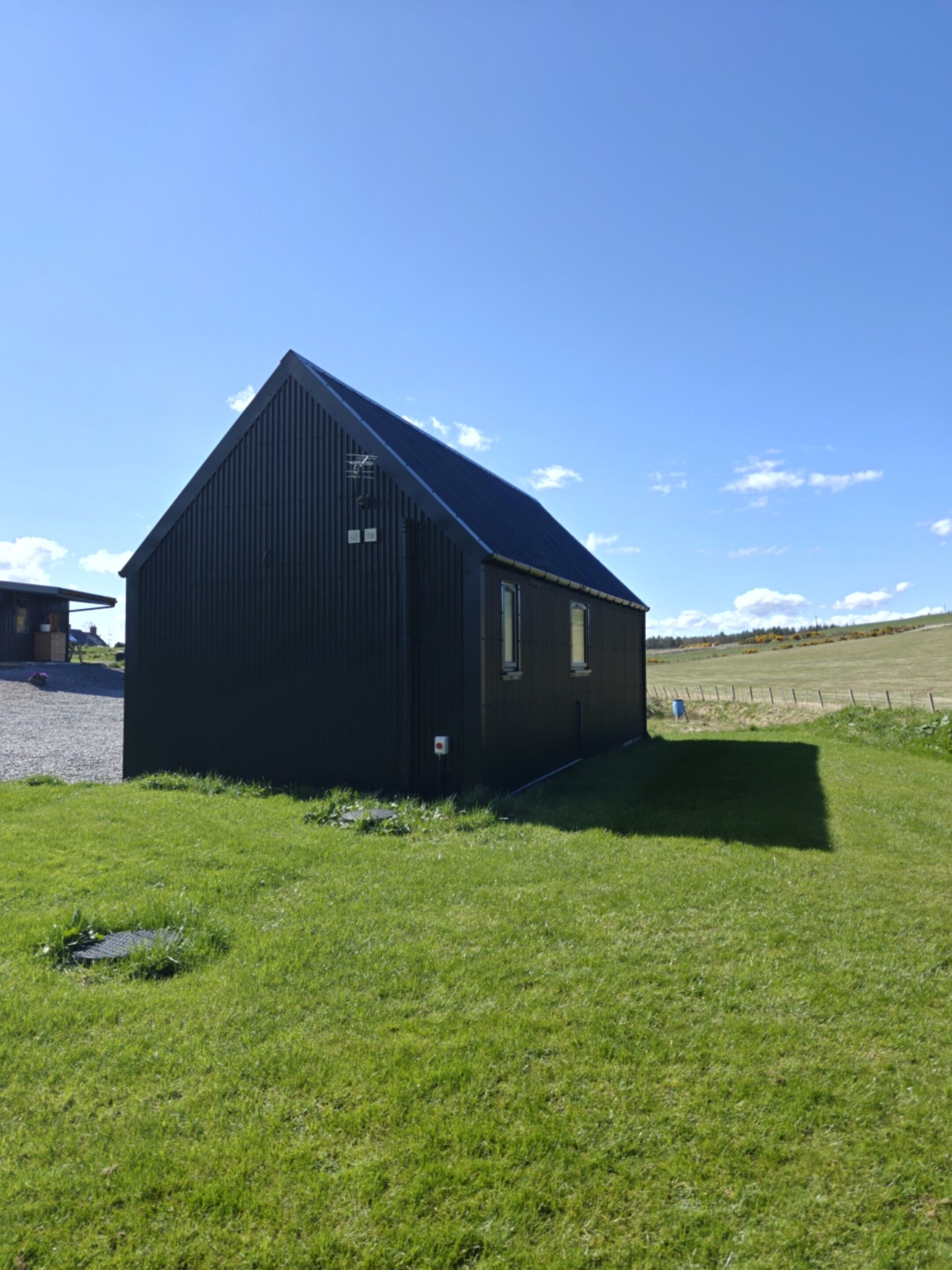 A black building with a sloped roof, a small window, and a door, situated on a grassy field with a clear sky.