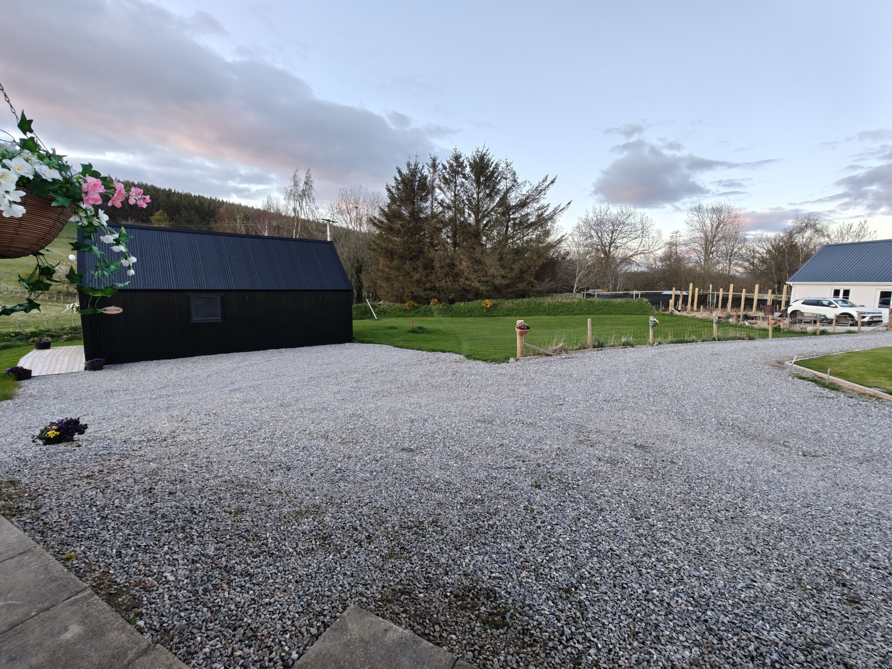A gravel driveway leading to a black building with a small window, surrounded by a fenced area with a grassy field and trees.
