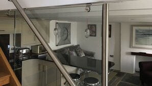Une cuisine moderne avec un escalier en verre, un coin repas avec une table en verre et un salon avec un canapé et une horloge au mur.