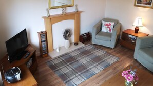 A living room with a fireplace, a television, a sofa, a coffee table, and a lamp. - Newholme Self-catering Bungalow (Pitlochry)