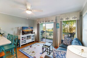 Une pièce dotée d’un ventilateur de plafond, d’un téléviseur posé sur un meuble blanc, d’un canapé agrémenté de coussins à motifs, d’une petite table et offrant une vue sur l’extérieur grâce à une porte coulissante.