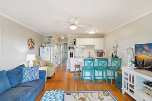 Un espace de vie compact comprenant un coin cuisine, un coin repas avec des chaises turquoise, un canapé bleu agrémenté de coussins à motifs et un téléviseur posé sur un meuble blanc.