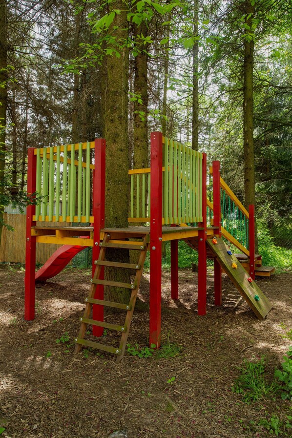 A wooden play structure with a ladder and slides in a wooded area.