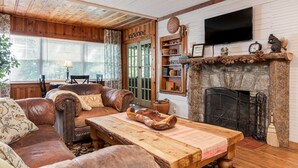 Un salon rustique avec un plafond en bois, un canapé en cuir, une table basse en bois et une cheminée surmontée d’une hotte en pierre.