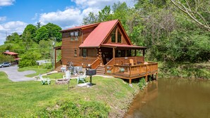 Une cabane en rondins avec un porche, une petite terrasse et une cour verdoyante aménagée avec des meubles d’extérieur.