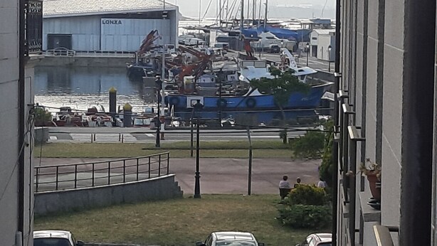 Vista de un puerto deportivo con barcos amarrados, un edificio con el rótulo "LONXIA" y un estacionamiento con coches.